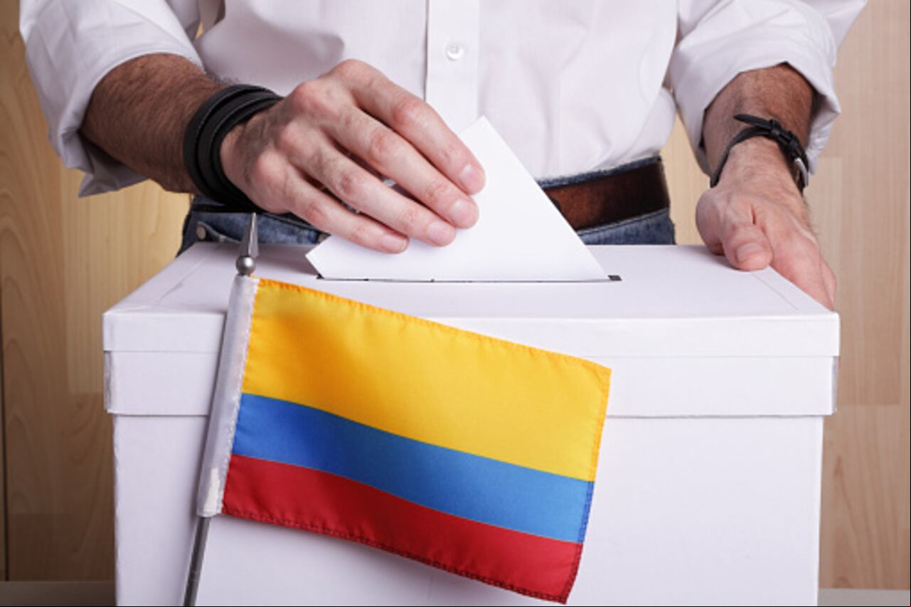 A man inserting a ballot to a ballot box. Colombian flag in front of it.