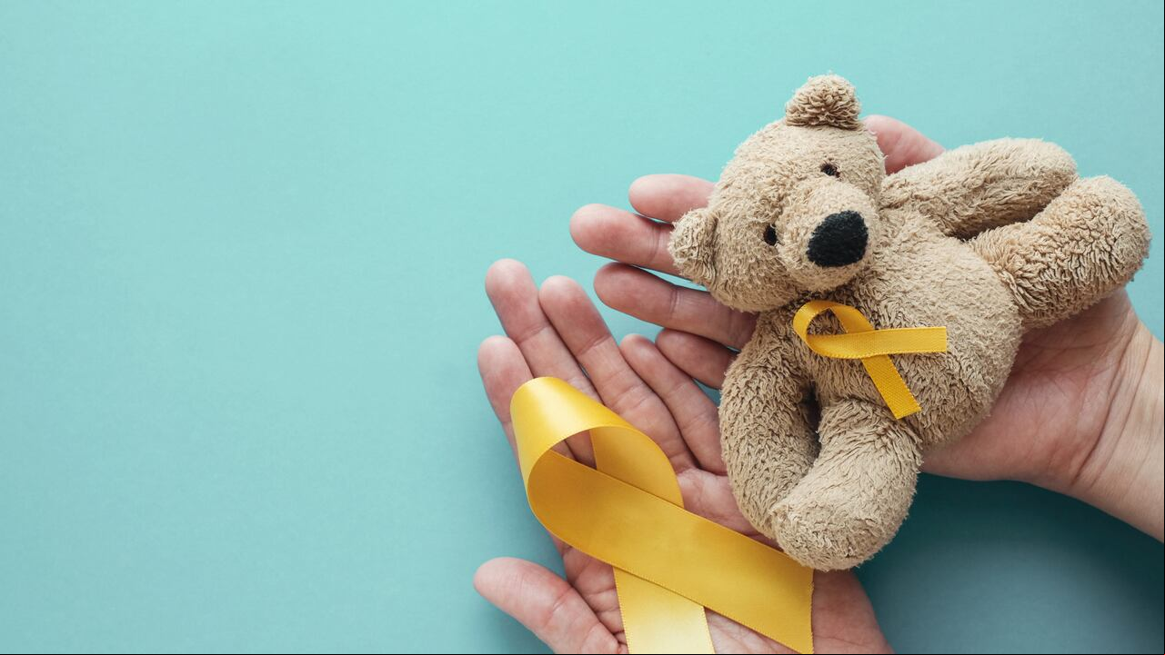 Manos sosteniendo a los niños oso pardo de juguete suave con cinta de oro amarillo, conciencia del cáncer infantil